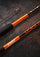 Close-up of an orange and black salmon fishing rod with white 'ANIRA' and 'SALMON' branding, laid diagonally on a dark textured surface with scattered orange flecks.