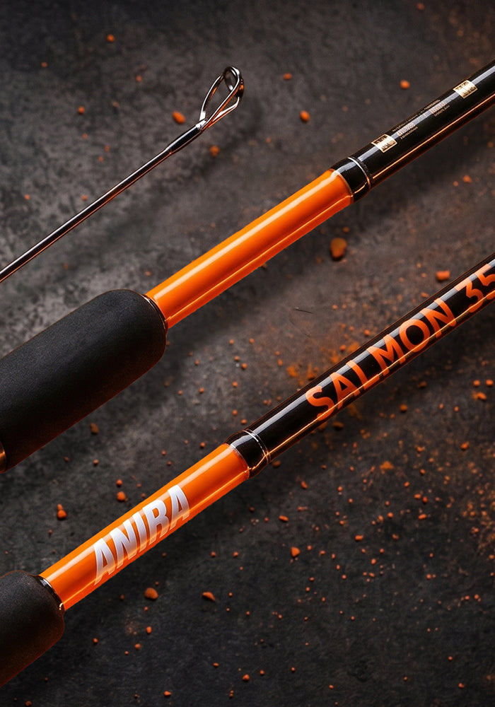 Close-up of an orange and black salmon fishing rod with white 'ANIRA' and 'SALMON' branding, laid diagonally on a dark textured surface with scattered orange flecks.