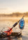 A close-up action shot of a fishing lure splashing into water at sunset. The lure has a blue and silver spinner blade with red and orange skirt strands, set against a blurred golden lake background.