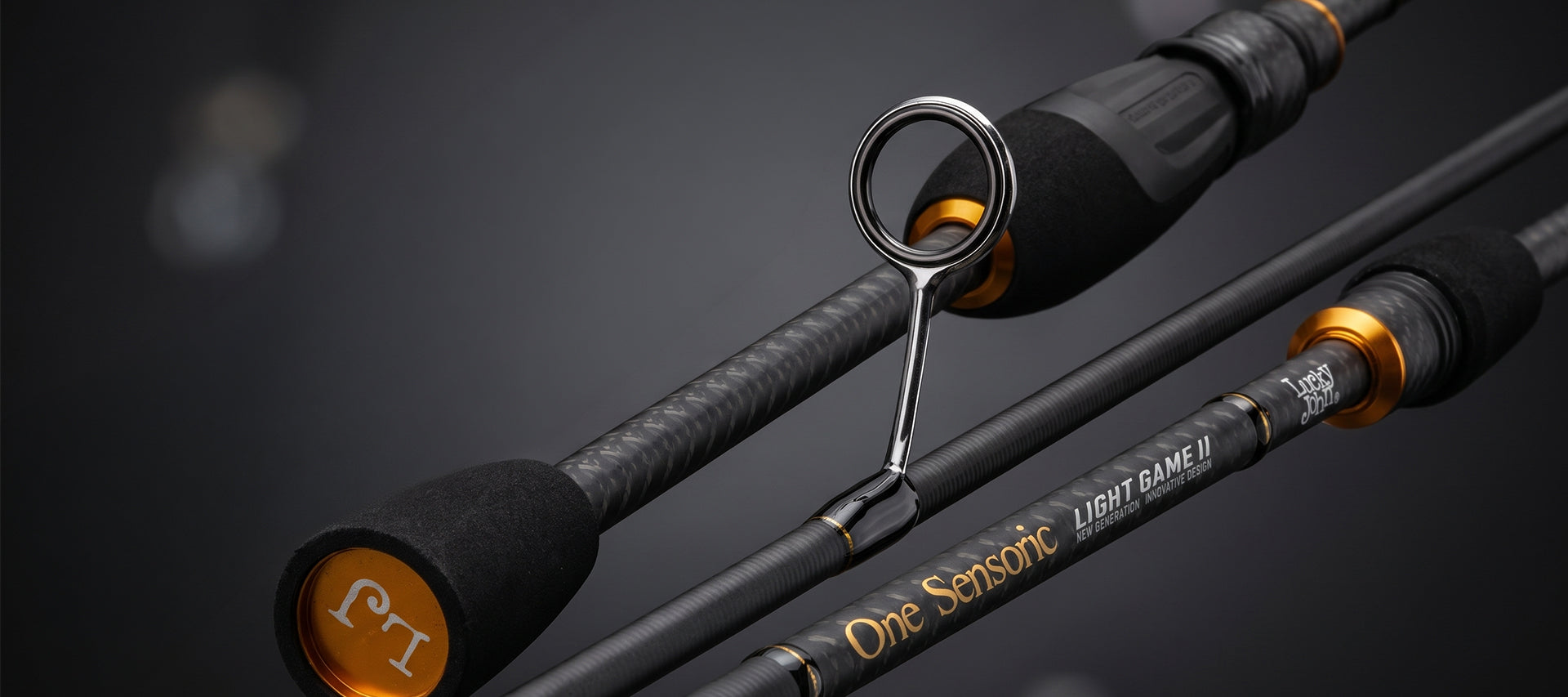 Close-up of a black and gray carbon-fiber fishing rod with gold accents and a silver line guide, shown against a dark blurred background. The barrel is labeled 'One Sensonic Light Game II'.