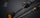 Close-up of a black and gray carbon-fiber fishing rod with gold accents and a silver line guide, shown against a dark blurred background. The barrel is labeled 'One Sensonic Light Game II'.
