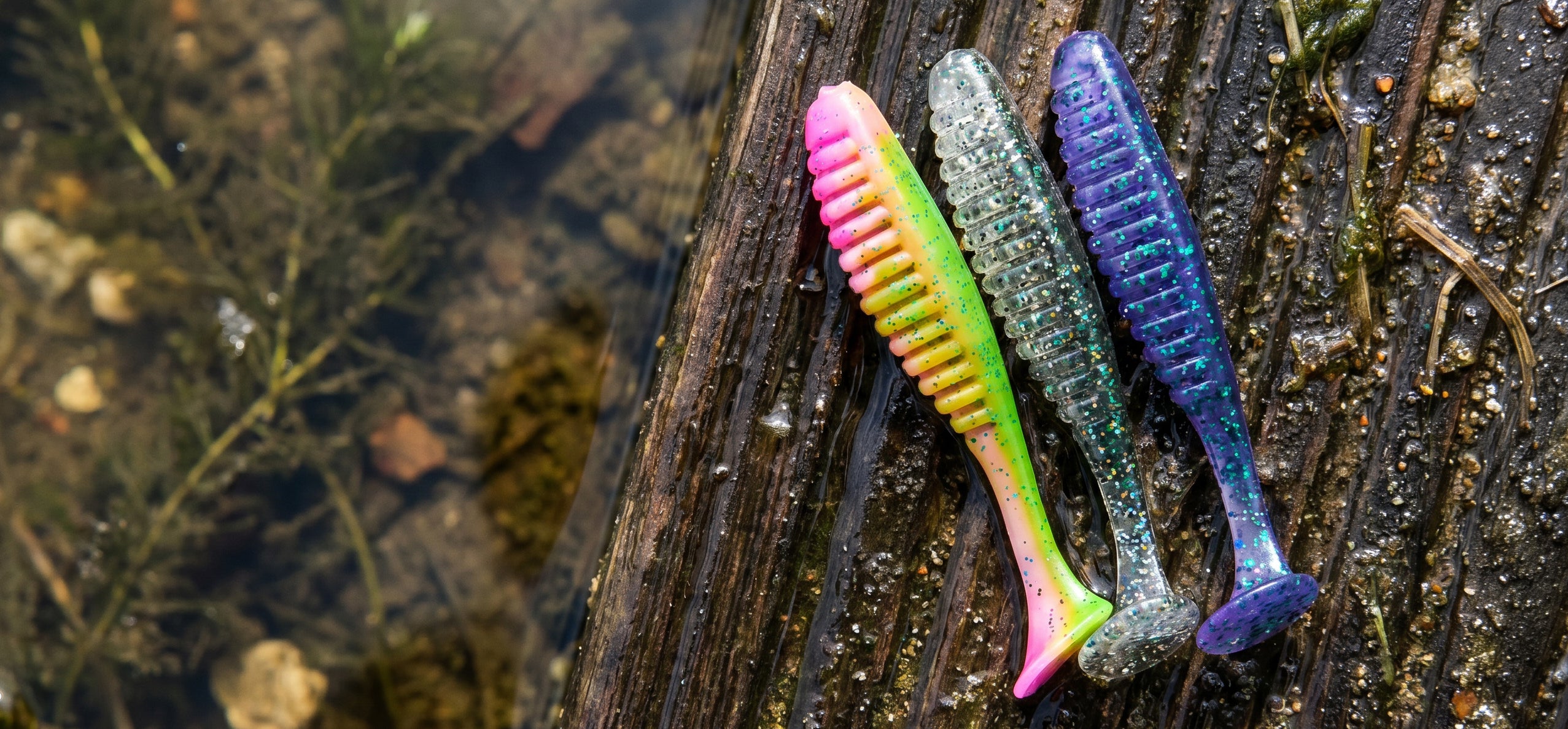 Three soft plastic fishing lures with ribbed bodies and bristles, resting on a wet dark wood log in shallow water. The lures have a gradient of neon pink, yellow, and green on one, a glittery light blue on the middle, and a glittery purple-blue on the right.