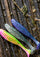 Three soft plastic fishing lures with ribbed bodies and bristles, shown on wet dark wood. The lures are in pink and yellow, green and silver, and blue and purple, all with glitter.