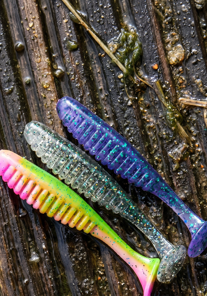 Three soft plastic fishing lures with ribbed bodies and bristles, shown on wet dark wood. The lures are in pink and yellow, green and silver, and blue and purple, all with glitter.