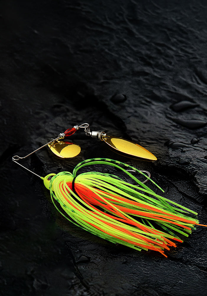 A fishing lure with two gold spinner blades and a neon green, orange, and yellow skirt, displayed on a dark textured slate surface.