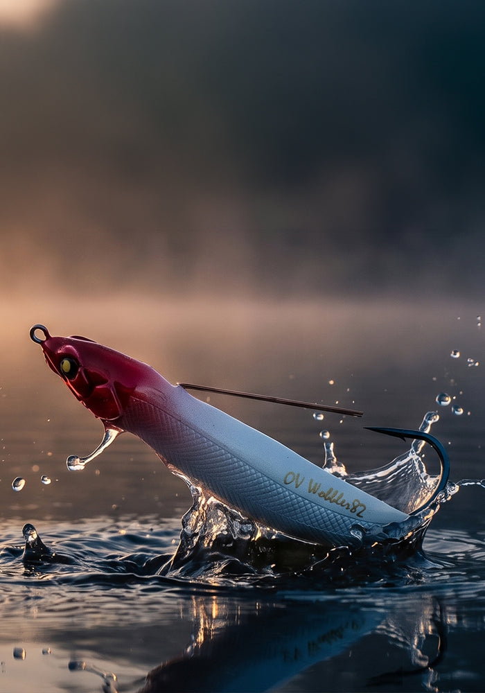 A fishing lure shaped like a small fish, shown mid-leap out of water with a splash. The lure has a red head, a silver body with a white lower section, and gold lettering that reads 'CV Kallefant'.