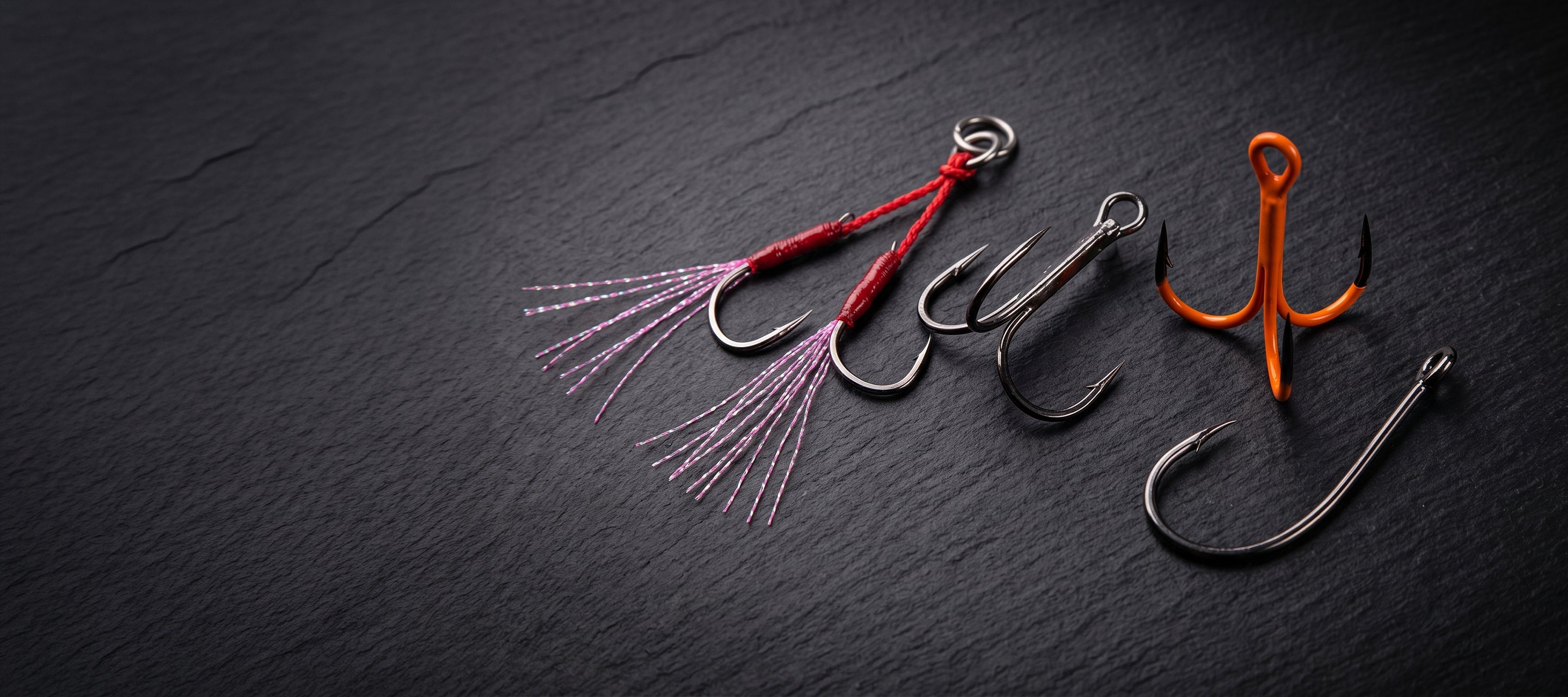 Three fishing hooks arranged on a dark textured slate background: one with a red body and pink bristles, one plain silver, and one with an orange top and silver hooks.