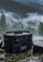 A black Lucky John waterproof bag with red trim sits in a shallow stream, with a misty forest and mountains in the background.