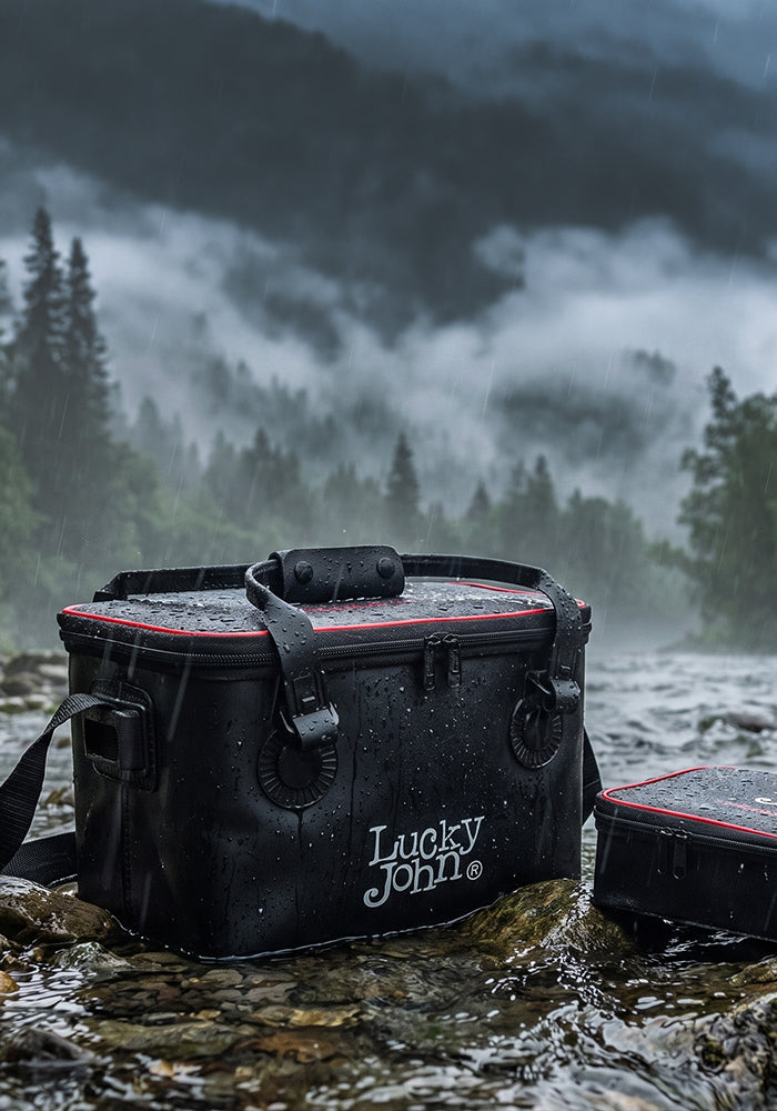 A black Lucky John waterproof bag with red trim sits in a shallow stream, with a misty forest and mountains in the background.