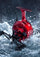 A glossy red spinning fishing reel with a black handle, emerging from splashing white water against a dark, dramatic background.