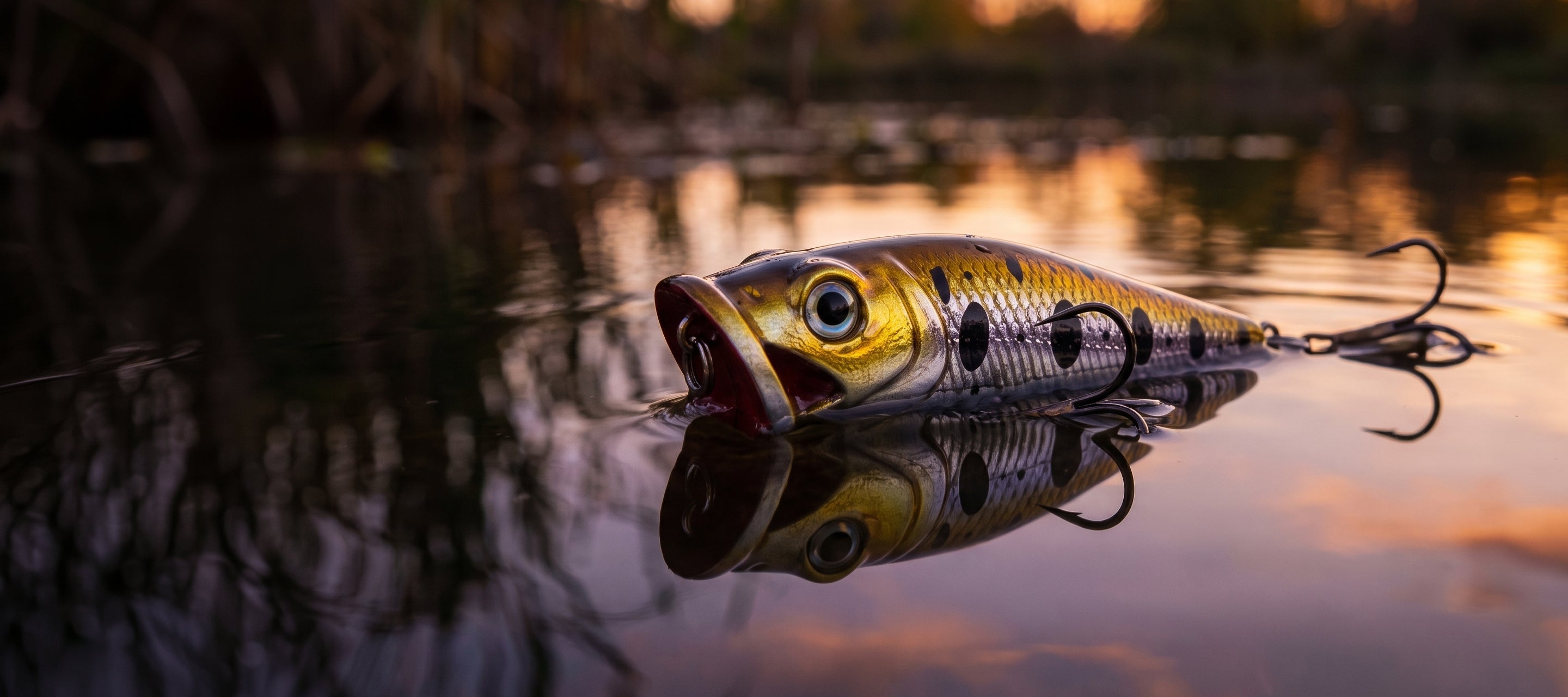 A realistic hard bait fishing lure shaped like a small fish, floating on calm water at sunset with its reflection visible. The lure has a yellow and black mottled body with a dark tail and treble hooks attached.